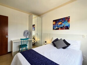 A bedroom with a bed, a desk with a chair, a standing fan, and a painting on the wall. - Residencial Dona Virginia (Canela)