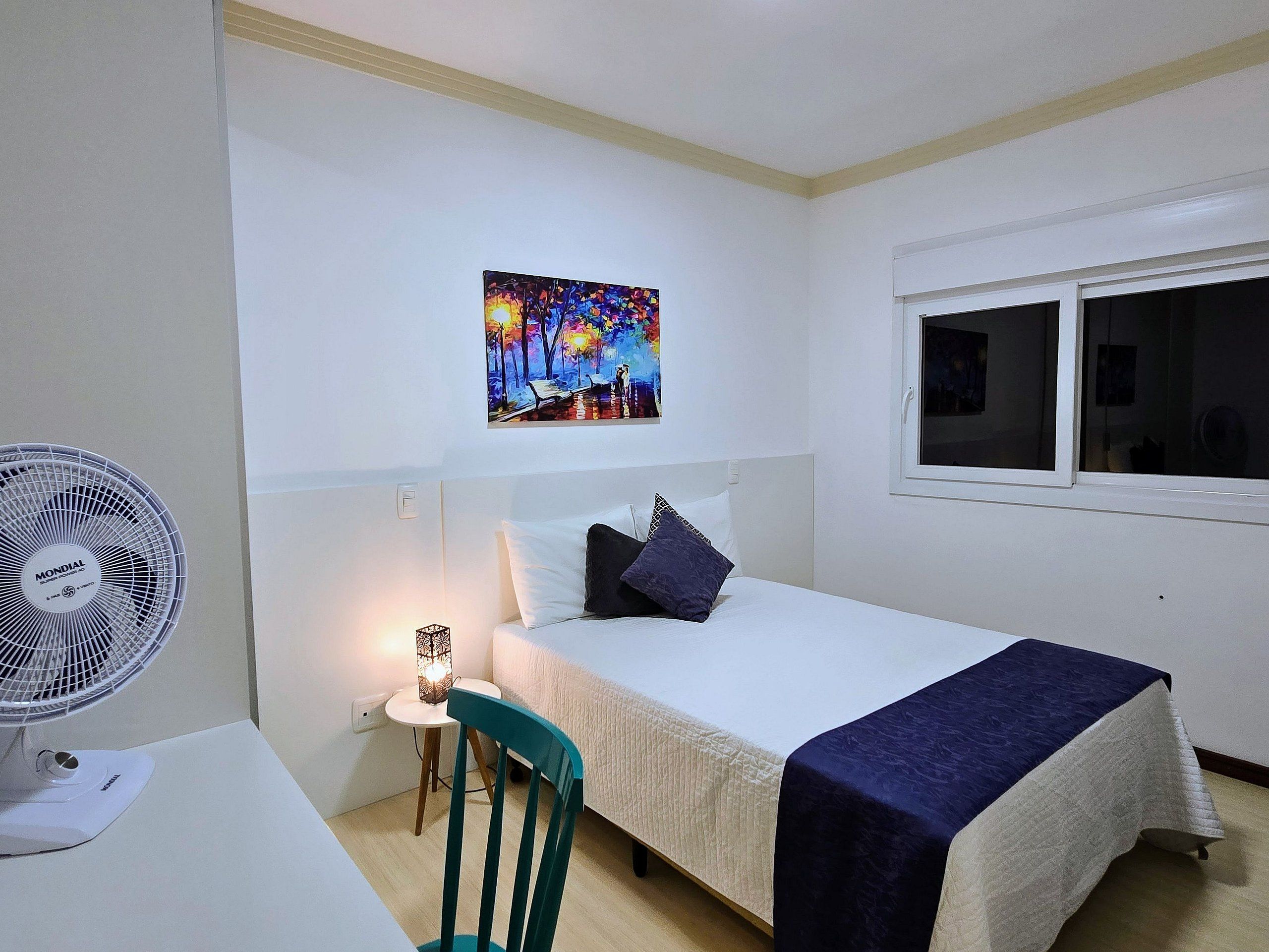 Un cuarto de hotel con cama, escritorio con ventilador, silla, mesita de noche y un cuadro en la pared.
