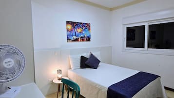 Un cuarto de hotel con cama, escritorio con ventilador, silla, mesita de noche y un cuadro en la pared.