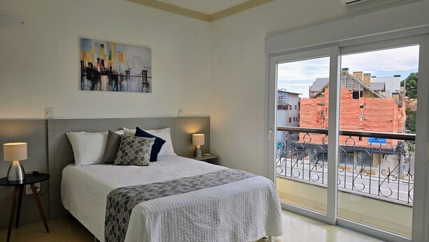 A bedroom with a bed, two bedside tables, a nightstand, a window with a view of buildings, and an air conditioning unit. - Residencial Dona Virginia (Canela)
