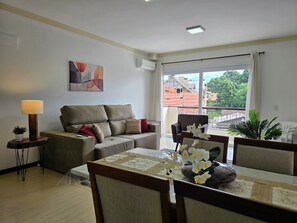 A living room with a sofa, a dining table with chairs, a lamp, and a window with a view of buildings and trees.
