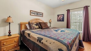 Une chambre avec un lit en bois, une table de chevet, une lampe, un ventilateur de plafond, une fenêtre avec des rideaux et un tableau accroché au mur.