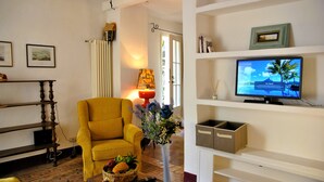 Een woonkamer met een gele fauteuil, een vaas met bloemen, een televisie op een dressoir en een boekenkast.