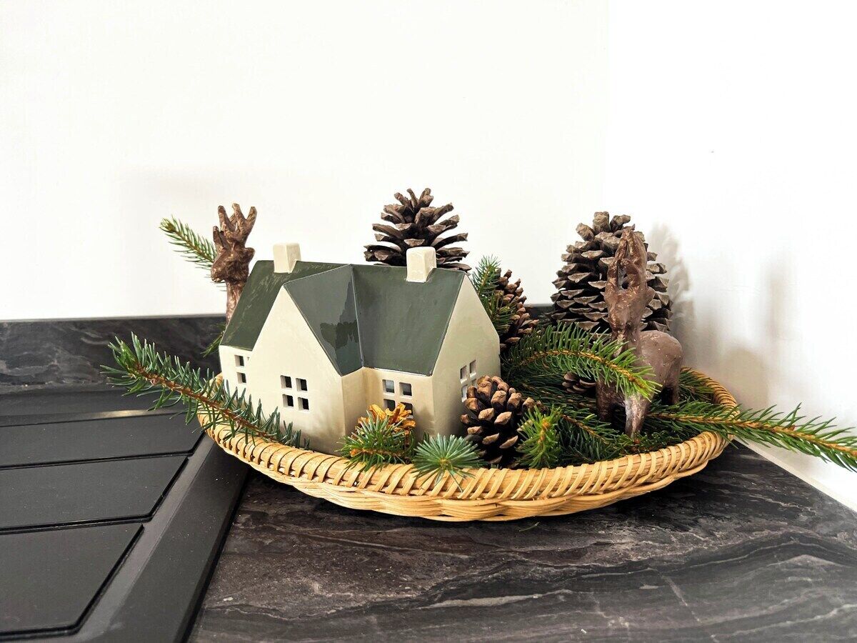 A small model house with a green roof and chimney, surrounded by pine cones and greenery on a dark surface.