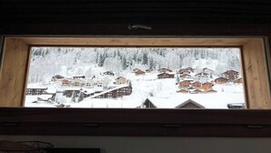 Une fenêtre donnant sur un village enneigé, avec des maisons en bois et une colline boisée.
