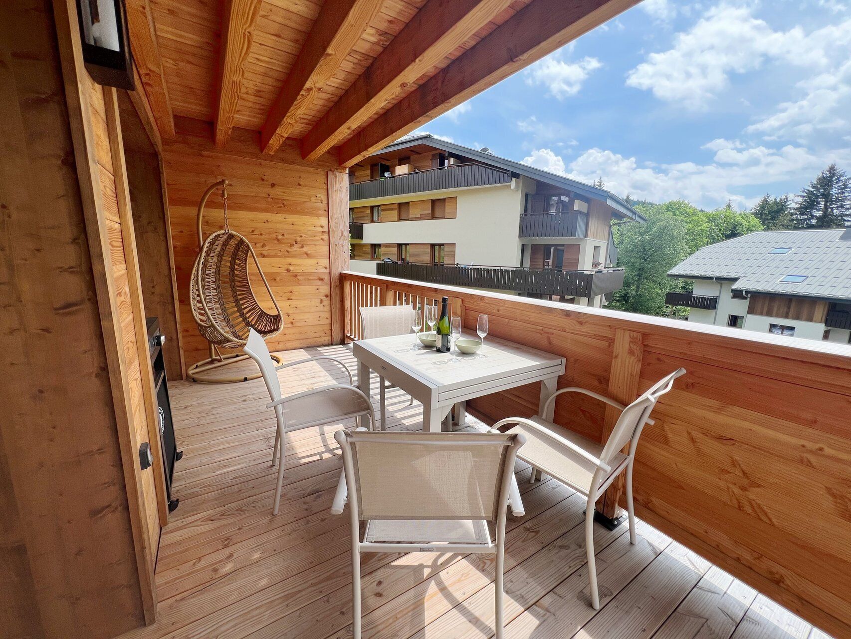 Ein Holzbalkon mit Tisch und Stühlen, einem Hängesessel und Blick auf angrenzende Gebäude.