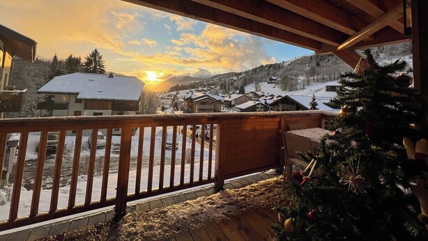 Un balcon en bois offrant une vue sur un village enneigé et des montagnes au coucher du soleil.
