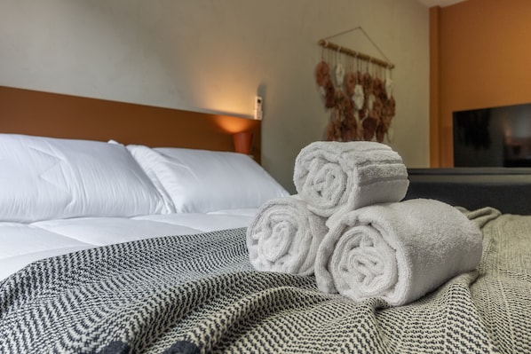 A neatly made bed with white linens, a patterned blanket, and rolled towels on a textured bedspread.