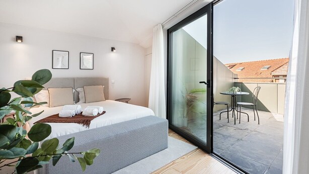 A modern bedroom with a large bed, a small table, and a sliding glass door leading to an outdoor patio. - GuestReady - Porto's Modern Comfort (Porto)