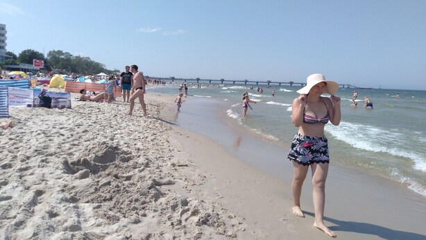 Une plage où l’on voit des gens se promener et nager, une jetée en arrière-plan et des parasols de plage.