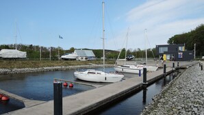 Un port de plaisance avec plusieurs bateaux amarrés, un petit bâtiment et une jetée s’avançant dans une eau calme.