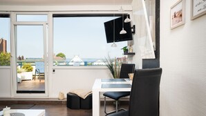 Een moderne woonkamer met een grote schuifpui naar een balkon, een eethoek met tafel en stoelen, en uitzicht op zee en gebouwen.