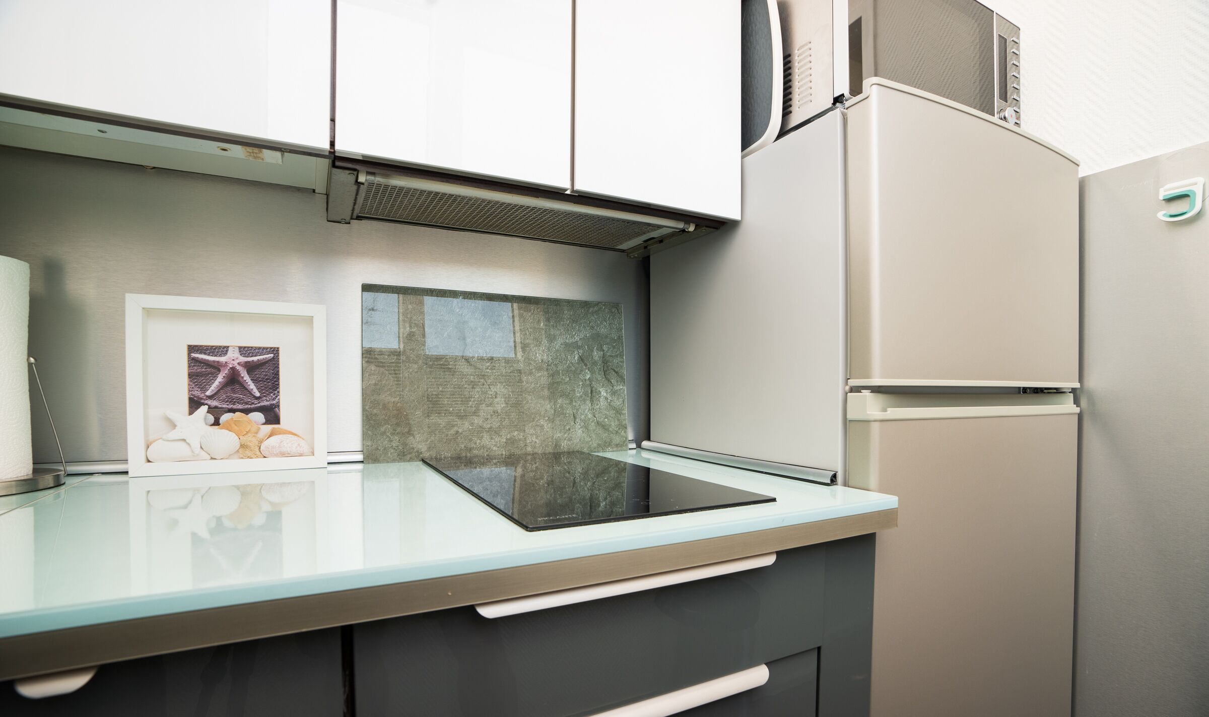 A modern kitchen with a glass countertop, stainless steel appliances, and a framed picture of seashells.