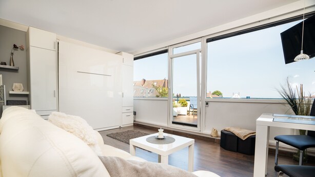 Een modern appartement met een grote schuifpui naar een balkon, witte meubels en uitzicht op de stad.