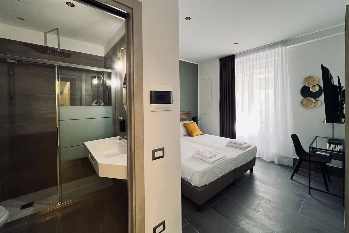 A modern hotel room with a walk-in closet, a large bed with a nightstand, a desk with a chair, and a bathroom with a glass shower enclosure.