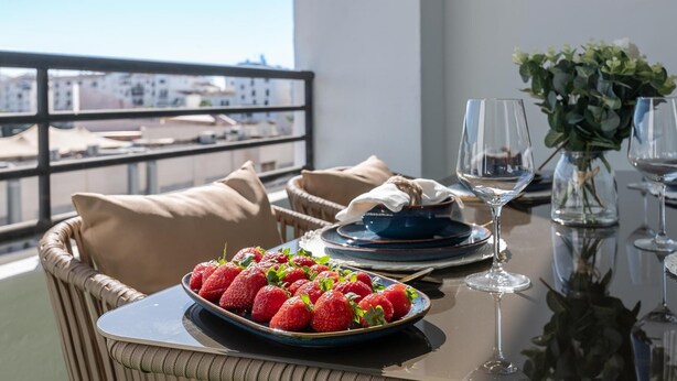 A dining table with a plate of strawberries, a bowl, and wine glasses on a balcony with a view of buildings. - Puerto Banus Serenity Retreat (Marbella)