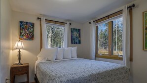 Ein Schlafzimmer mit einem Bett, einem Nachttisch, einer Lampe, zwei Fenstern mit Vorhängen und zwei Bildern an den Wänden.