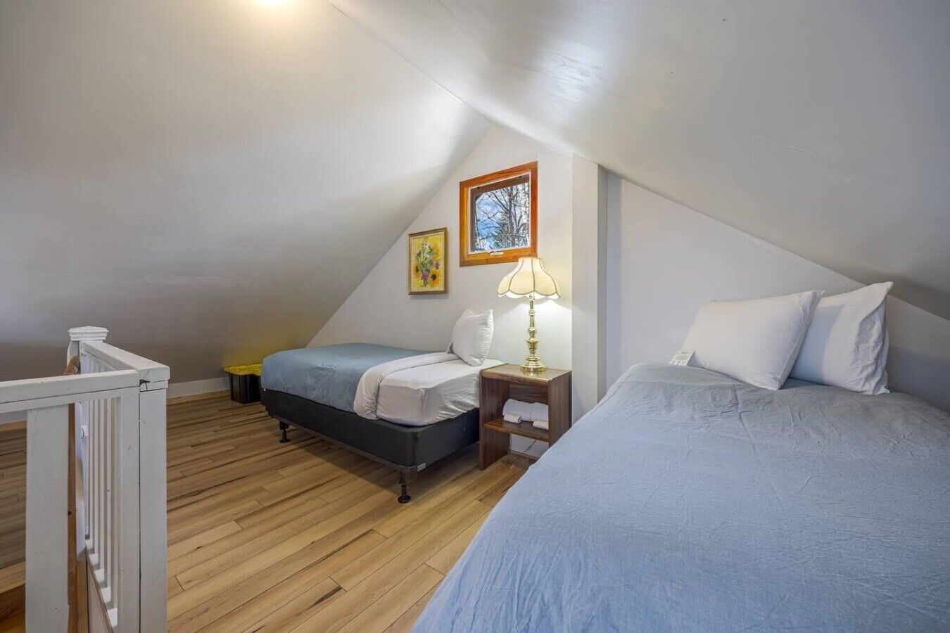Un dormitorio con dos camas, suelo de madera, una ventana con vistas y una mesita de noche pequeña con lámpara.