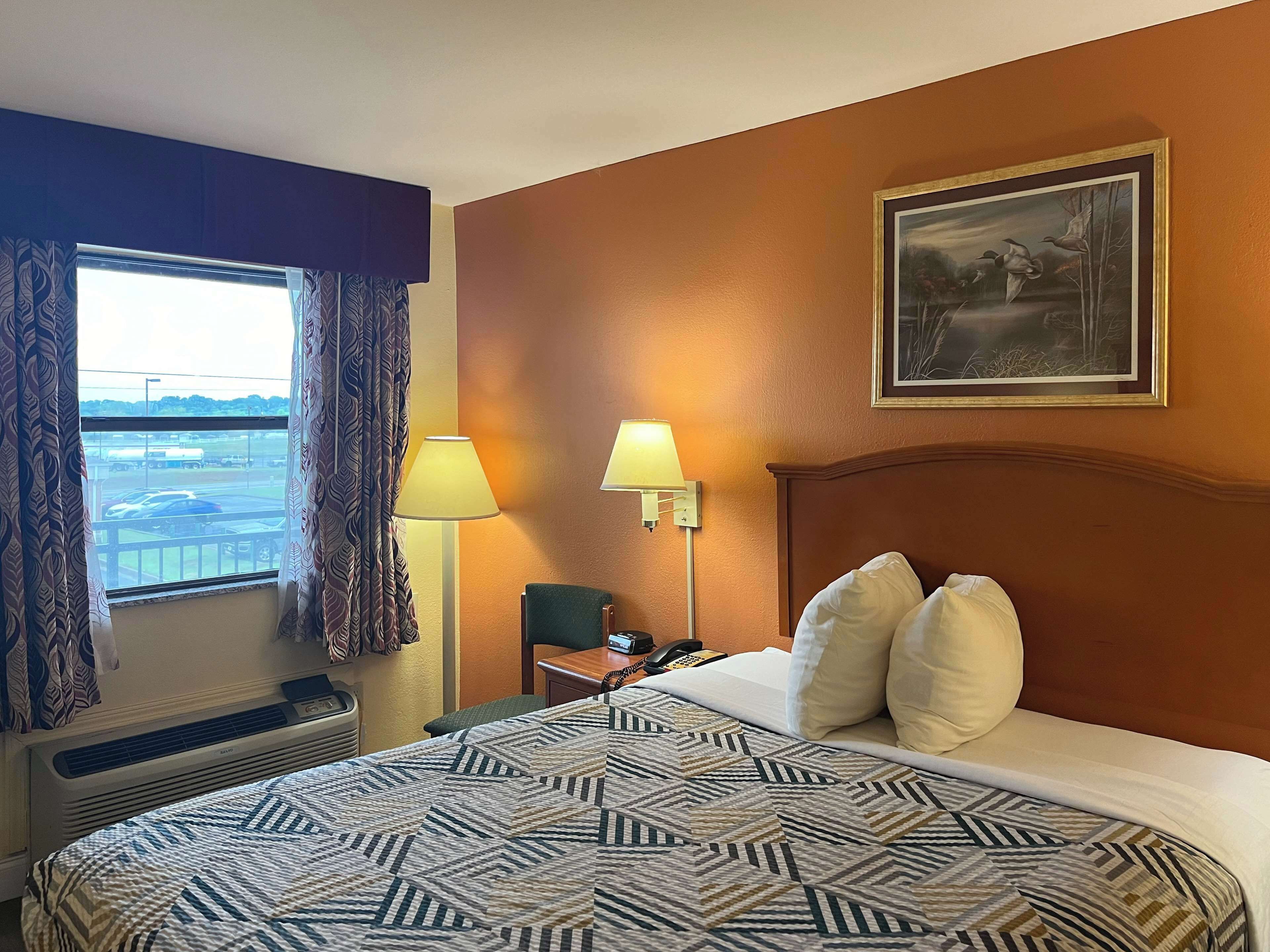 A hotel room with a large bed, two bedside lamps, a painting on the wall, and a view of the airport through the window.