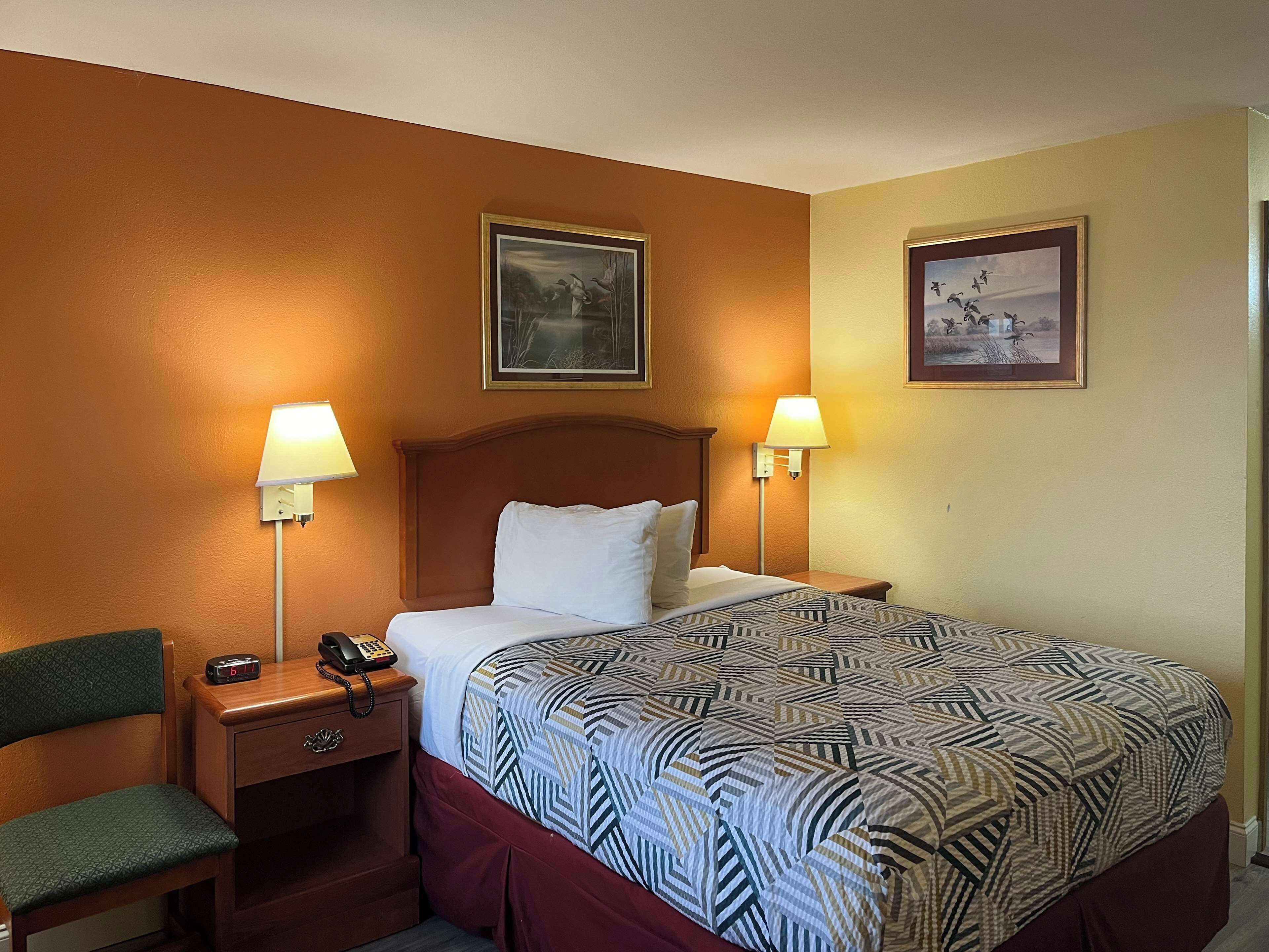 Quarto de hotel com uma cama grande, duas luminárias de cabeceira, mesa de cabeceira com telefone, uma cadeira e duas quadros nas paredes.