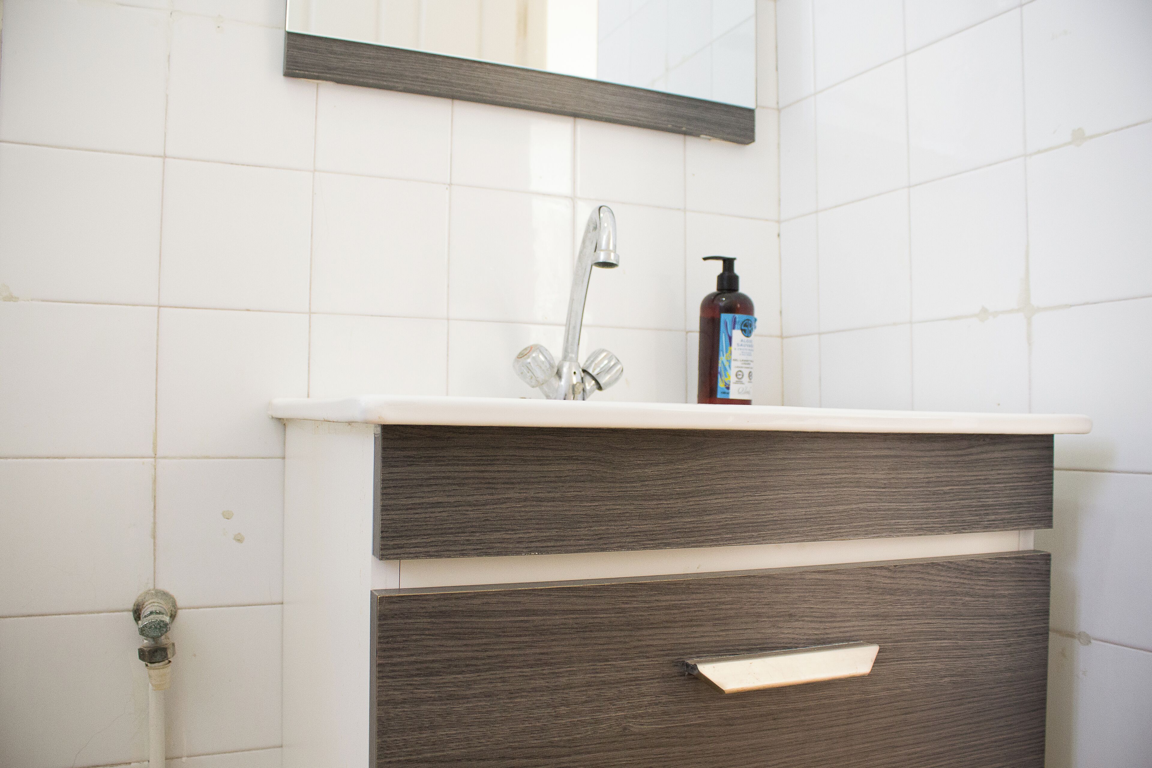 A bathroom with a white tiled wall, a wooden vanity with two drawers, a mirror, and a faucet.