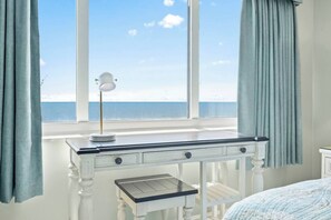 A bedroom with a desk by a window overlooking the sea, a lamp on the desk, and a bed with a blue blanket.