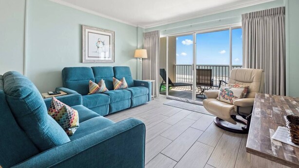 A living room with a sofa, armchair, and a table. There is a balcony with a view of the beach. - Oceanfront Delight - Indoor Pool, Sleeps 4 (North Myrtle Beach)