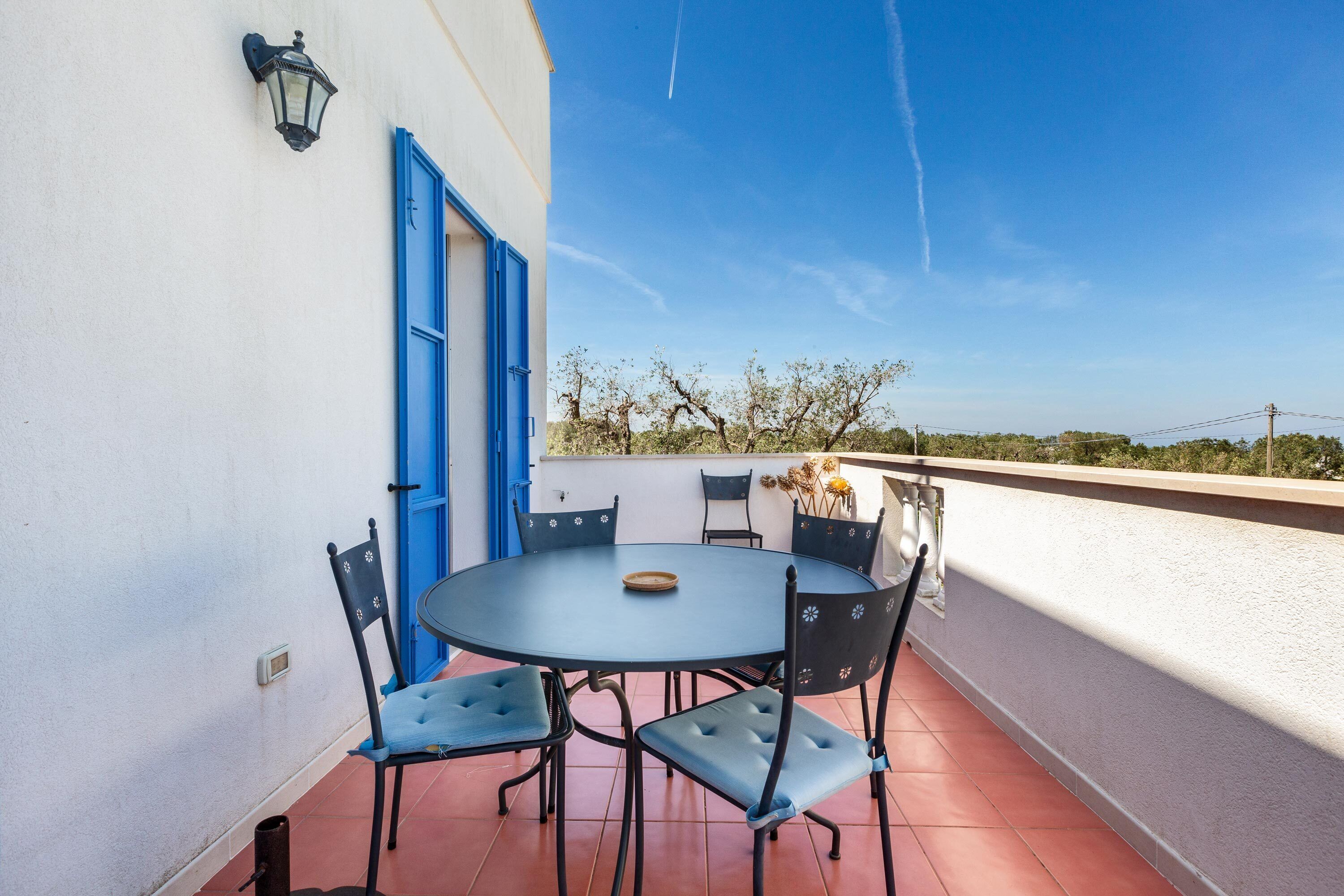 Een ronde tafel met stoelen op een balkon, met een blauwe deur en een wandlamp.