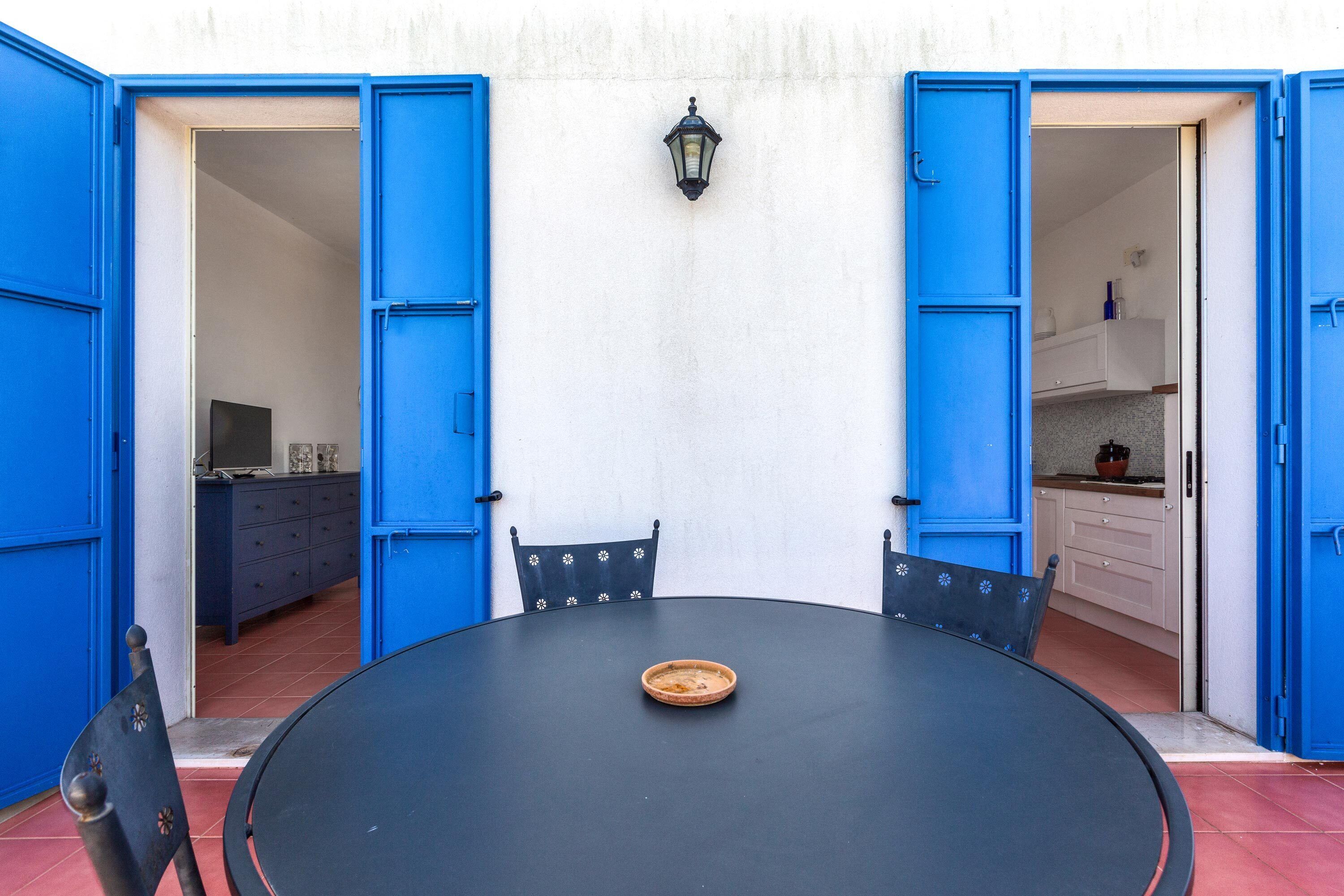 Una mesa redonda con un plato en el centro, acompañada por dos sillas, en una habitación con puertas azules y una lámpara en la pared.