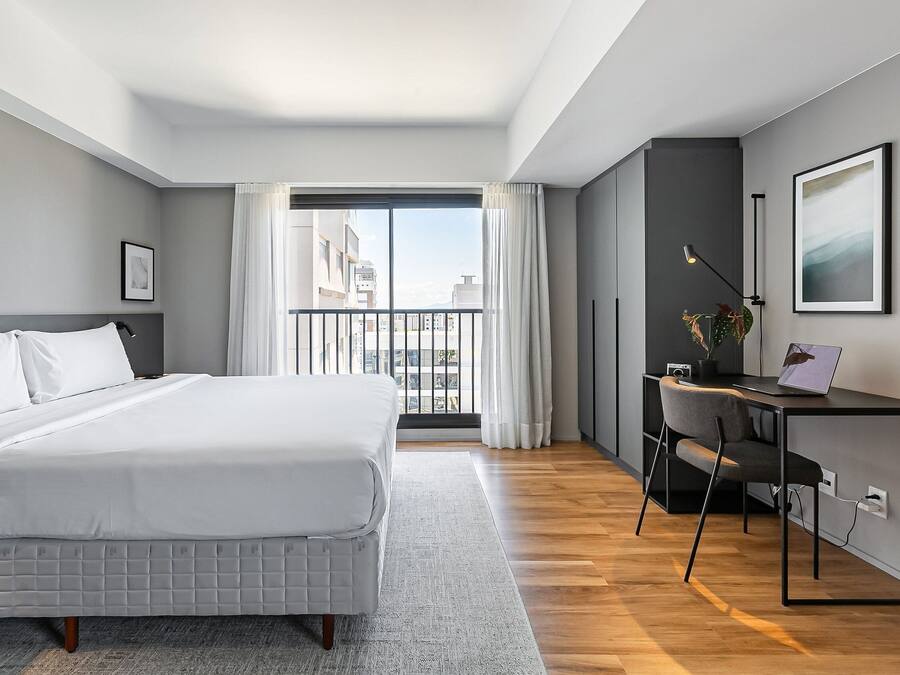 A modern bedroom with a bed, a desk, and a view of the city through a balcony.