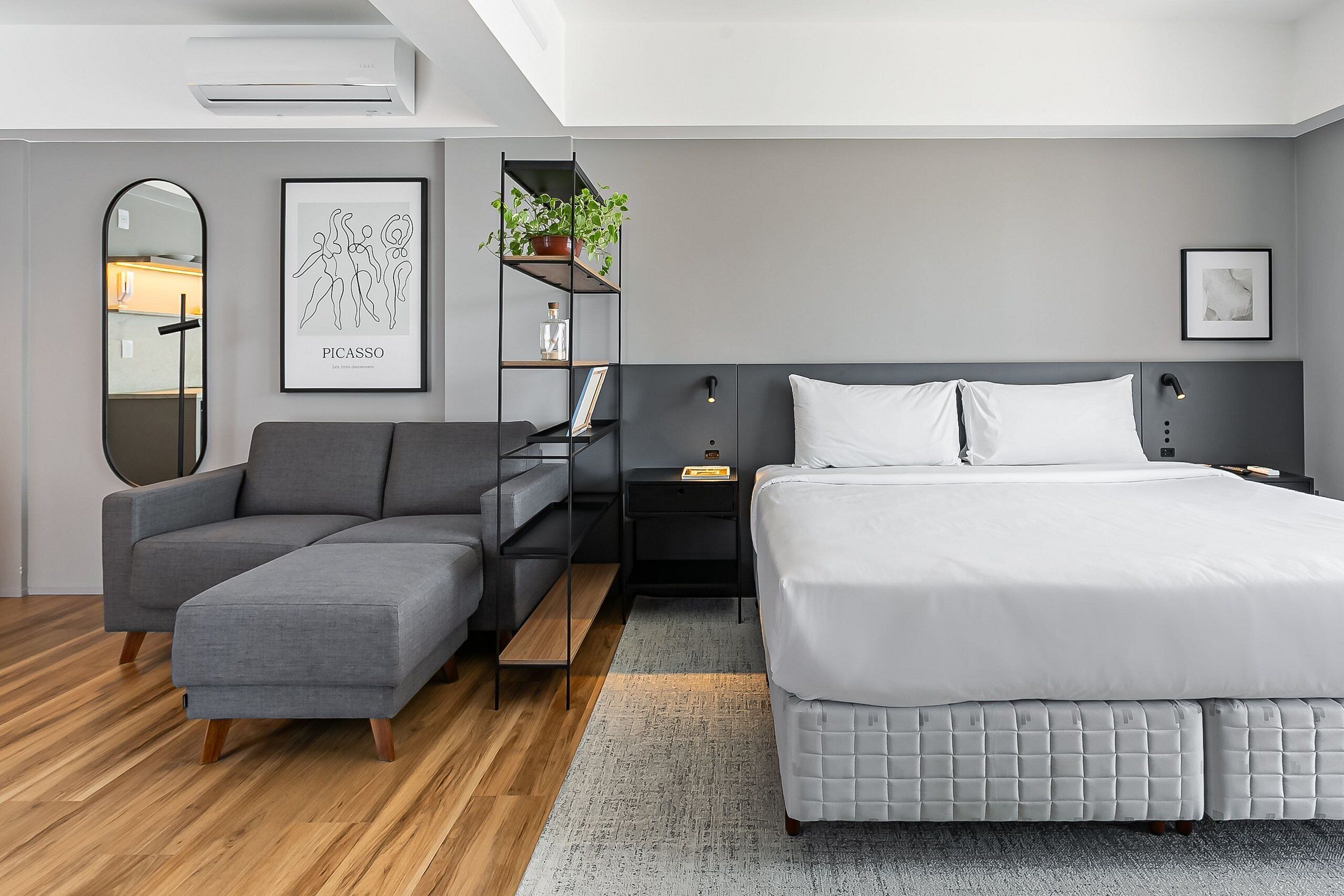 A modern bedroom with a grey sofa, a bed with white bedding, a nightstand, a bookshelf, and a mirror.