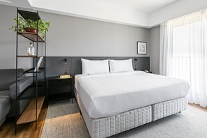 A modern bedroom with a large bed, a bedside table, a nightstand, a bookshelf, and a window with curtains. - Studios Rio Branco by Viva - Prédio moderno, aptos com cama king-size no centro de Floripa (Florianópolis)