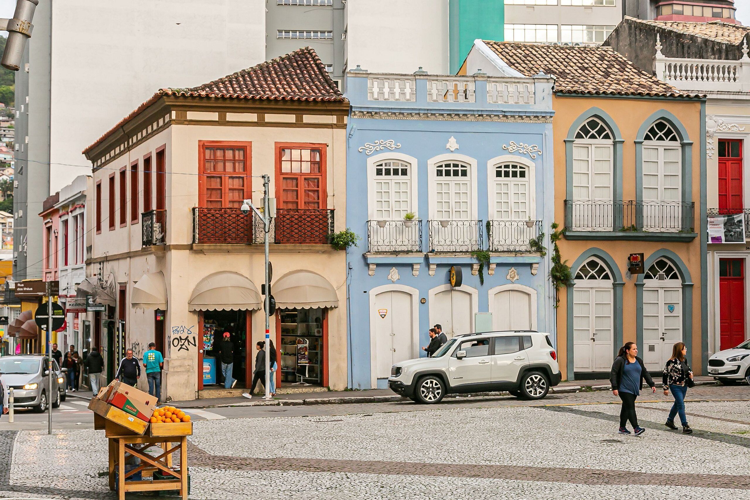 A street with colorful buildings, a white car parked on the side, and a fruit stand.