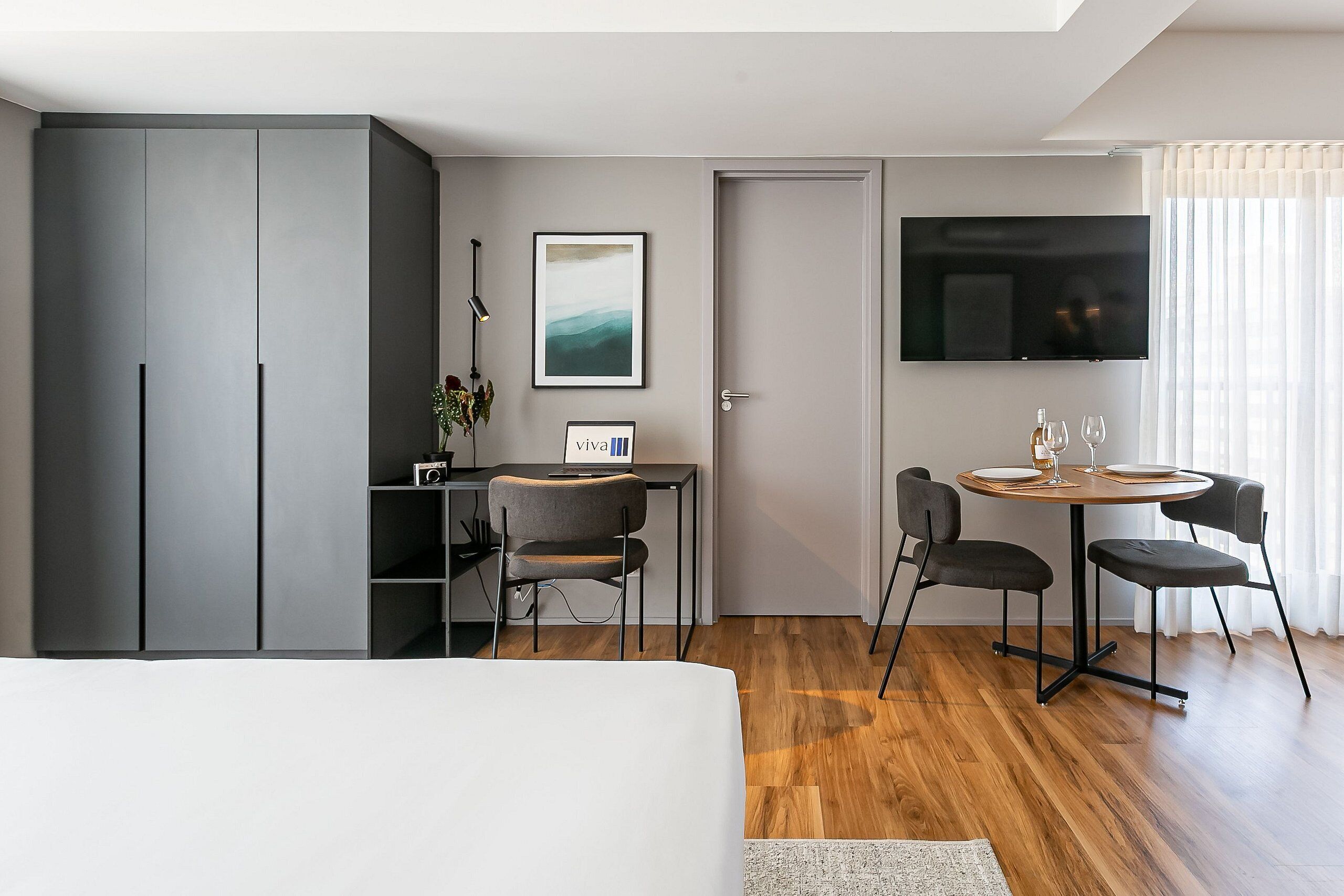 A modern hotel room with a bed, a desk with a chair, a TV, and a dining area with chairs and a table.
