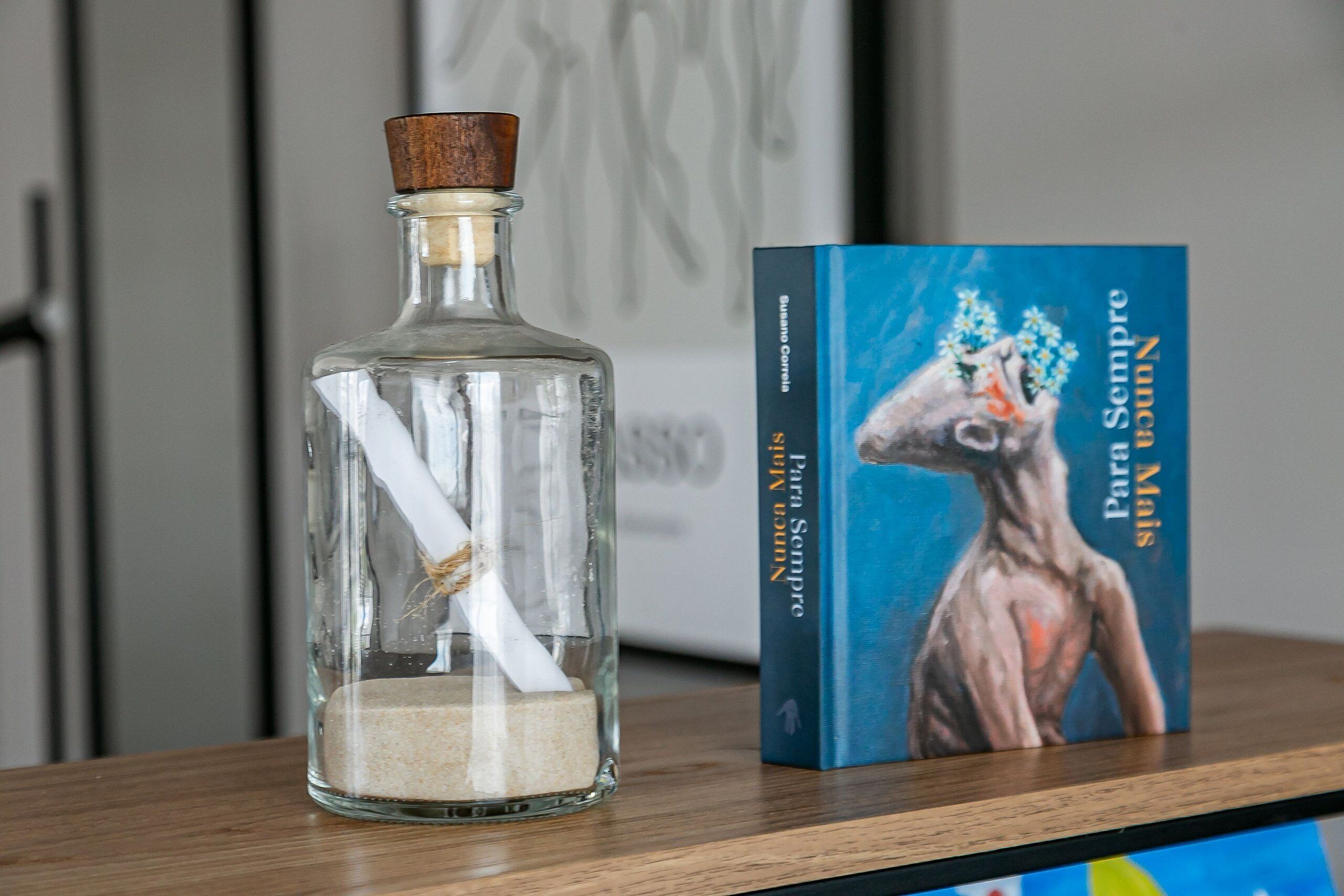 A clear glass bottle with a cork stopper and a white straw, placed on a wooden surface next to a book titled 'Nunca Sempre Para Sempre' by Susana Corrêa.