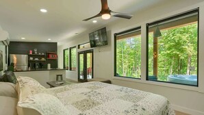 A modern bedroom with a large bed, a ceiling fan, a TV, and a view of a hot tub through large windows. - Love Shack cabin at Trickle Creek (Logan)