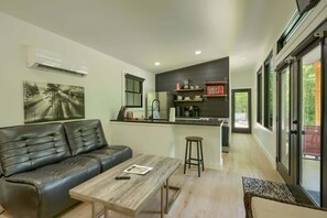 A modern living space with a sofa, a wooden coffee table, a kitchen area with a refrigerator, and a view through large windows.