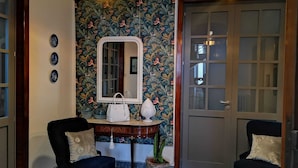 A room with a floral wallpaper, a mirror, two chairs, a small table with a white handbag, and a door with glass panels.
