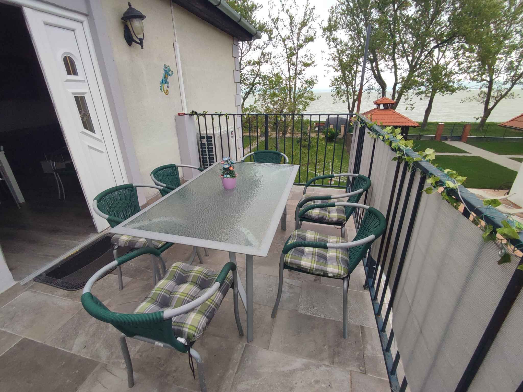Un patio al aire libre con una mesa de vidrio y sillas de mimbre, una puerta blanca y vistas a un cuerpo de agua.