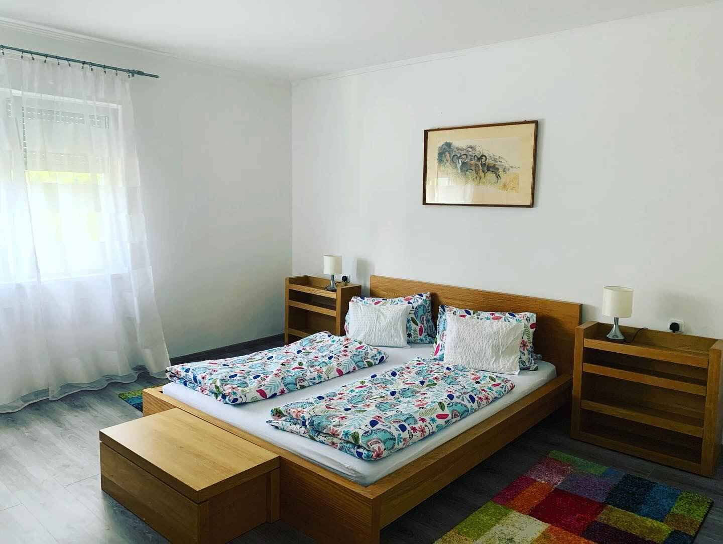 Un lit double avec une literie à motifs floraux, des tables de chevet avec des lampes, un banc en bois, un tapis coloré et un tableau encadré au mur.