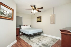 Een slaapkamer met een enkel bed, een plafondventilator, een ingelijst foto en een houten dressoir.