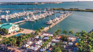 Un port de plaisance où sont amarrés de nombreux bateaux, un parking rempli de voitures et une plage bordée de palmiers et parsemée de tentes.