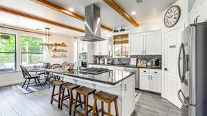 A modern kitchen with white cabinetry, a central island with stools, a stainless steel refrigerator, and a large window providing natural light. - Enchanting Clovis Haven w/ Hot Tub & Private Pool! (Clovis)