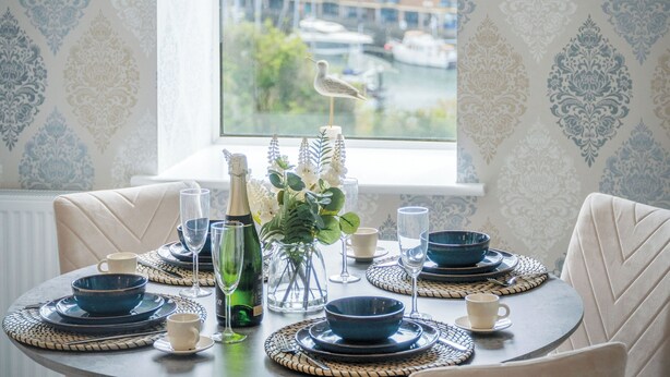 A dining table set with blue and white plates, bowls, and glasses, with a bottle of champagne and a vase of flowers. - Harbour View - 2 Bedroom Apartment - Milford Haven (Milford Haven)