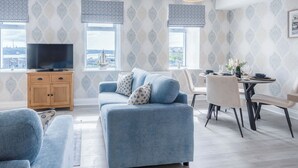 A living room with a blue sofa, a TV on a wooden cabinet, and a dining area with a table and chairs. - Harbour View - 2 Bedroom Apartment - Milford Haven (Milford Haven)