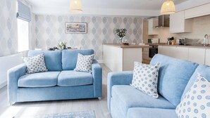 A modern living room with a blue sofa set, a kitchen area, and a dining table with a vase of flowers. - Harbour View - 2 Bedroom Apartment - Milford Haven (Milford Haven)