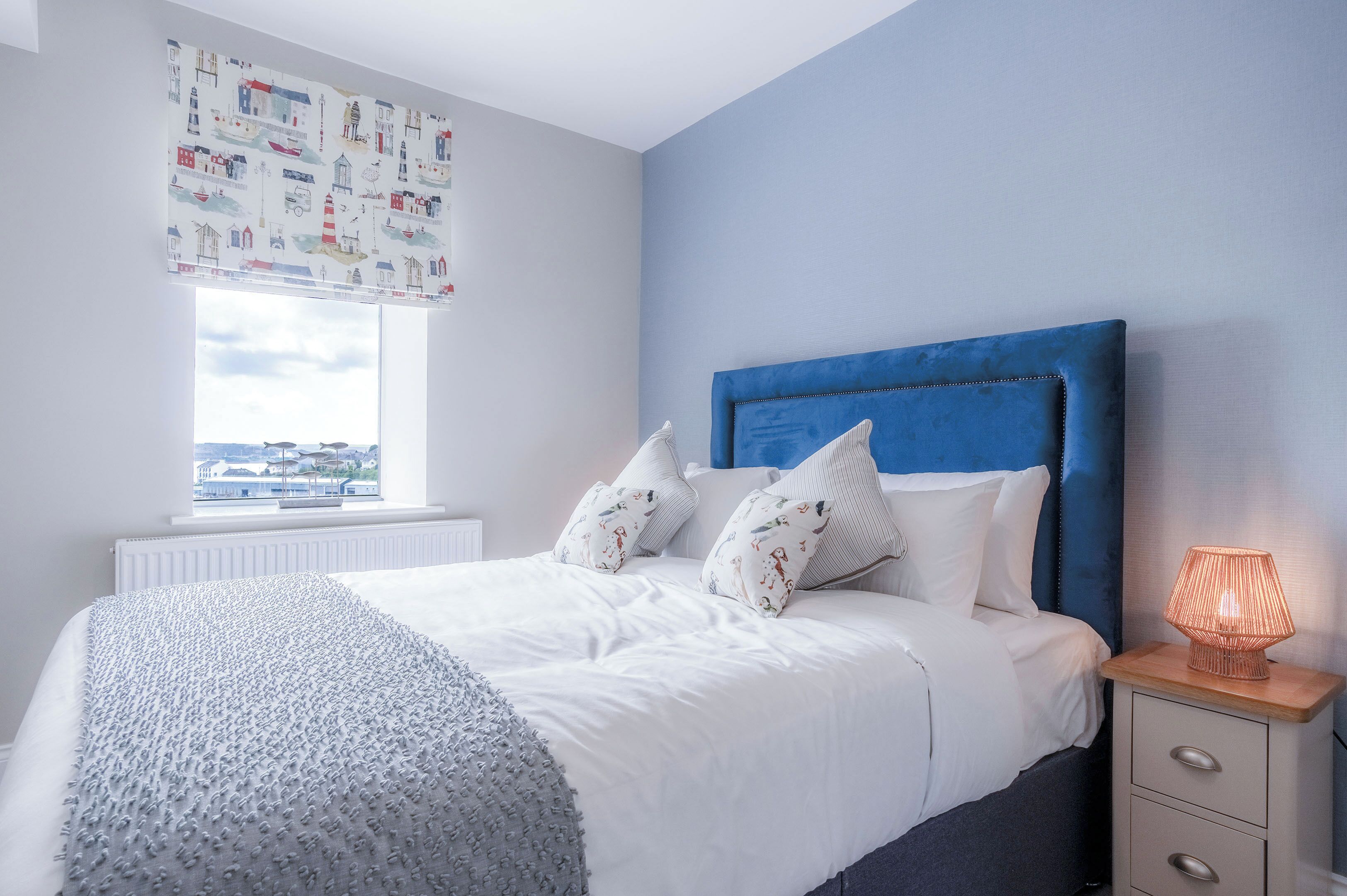 A bedroom with a bed, a nightstand, a lamp, a window, and a wall with a pattern.