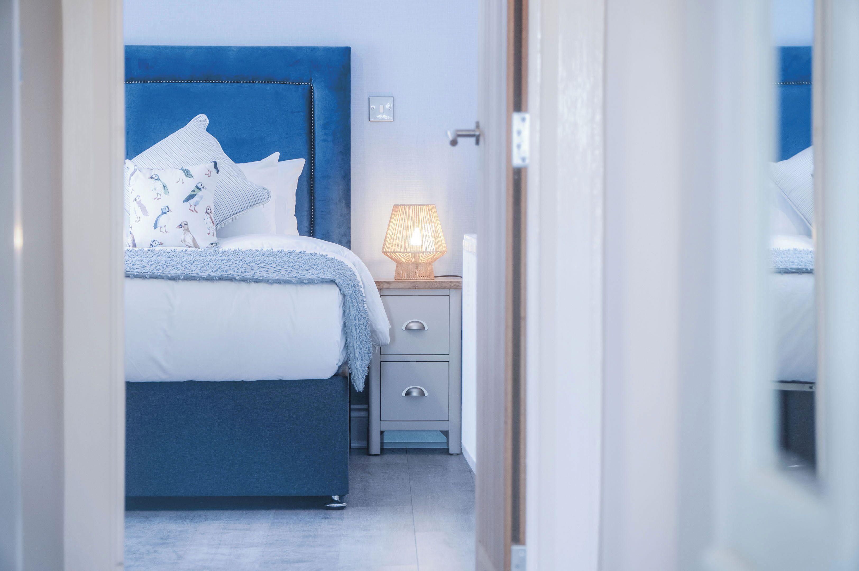 A bedroom with a blue headboard, white bedding, a bedside lamp, and a nightstand with drawers.