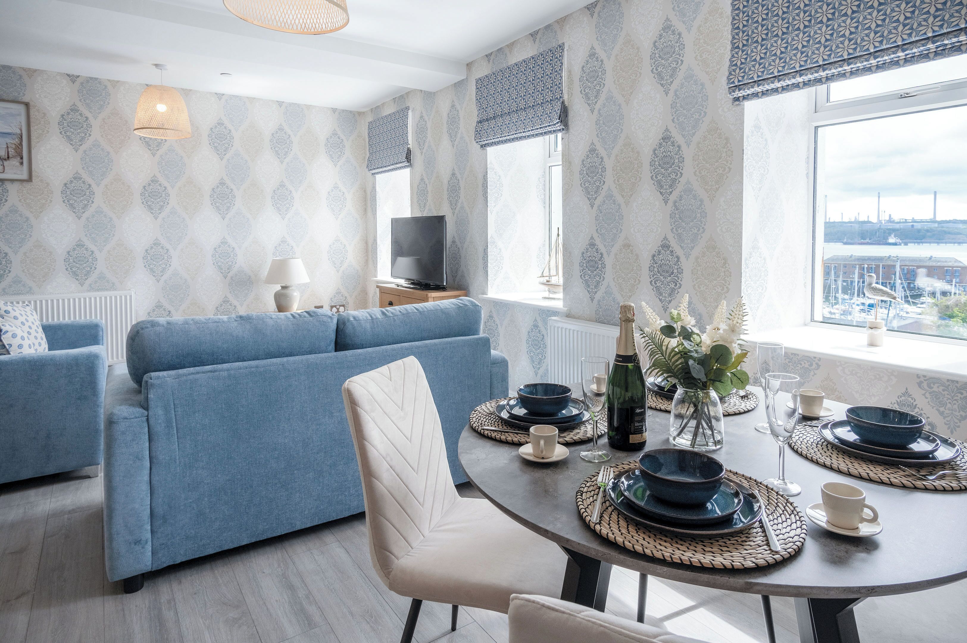 Una sala de estar moderna con un sofá azul, una mesa de comedor lista para cuatro comensales y un amplio ventanal con vistas a un paisaje urbano.