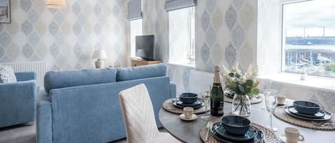 Una sala de estar moderna con un sofá azul, una mesa de comedor lista para cuatro comensales y un amplio ventanal con vistas a un paisaje urbano.